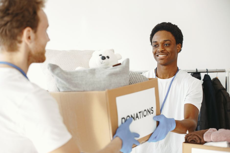 A young man with dark skin, wearing a white t-shirt and blue gloves, is smiling while handing over a cardboard box labeled 'DONATIONS' to another person with light skin and red hair, who is also weari