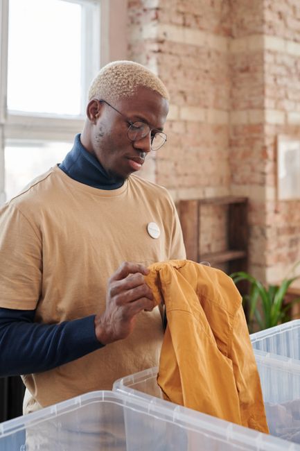 A young man with dark skin, wearing a white t-shirt and blue gloves, is smiling while handing over a cardboard box labeled 'DONATIONS' to another person with light skin and red hair, who is also weari