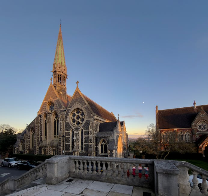 A detailed view of a historic church with Gothic-style architecture, featuring a tall spire, ornate stone carvings, pointed arched windows, and circular stained glass elements. The building’s exteri